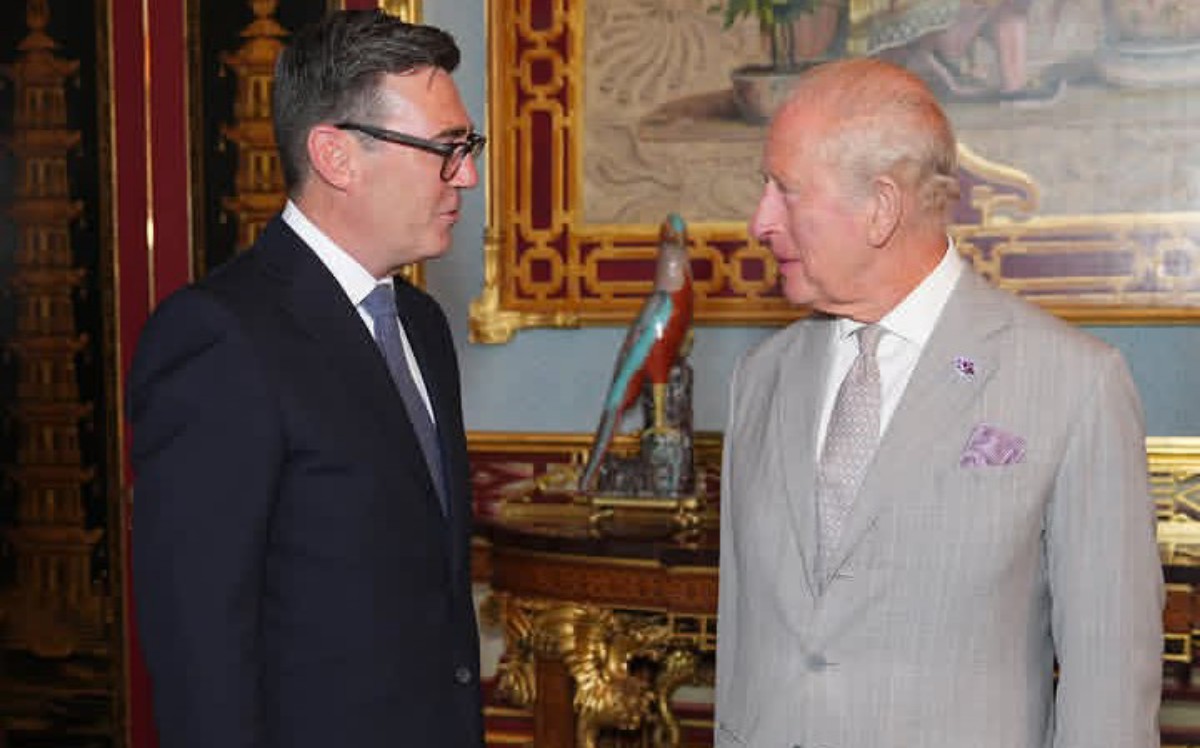 Two men in suits converse in an opulent, gold-accented room with a turquoise parrot statue on a pedestal between them.