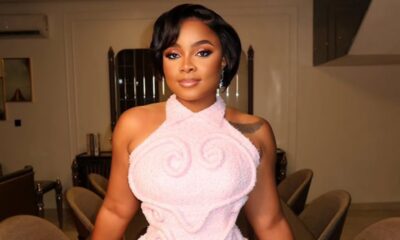 Woman in a pale pink halter dress posing in a beige room with chairs in the background, looking at the camera.