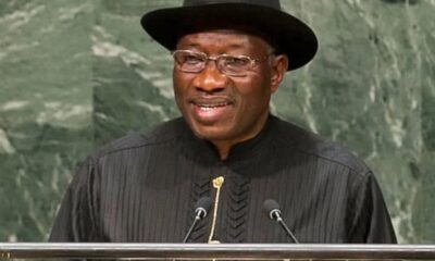 Man wearing a black hat and traditional black robe speaks at a podium with two microphones.