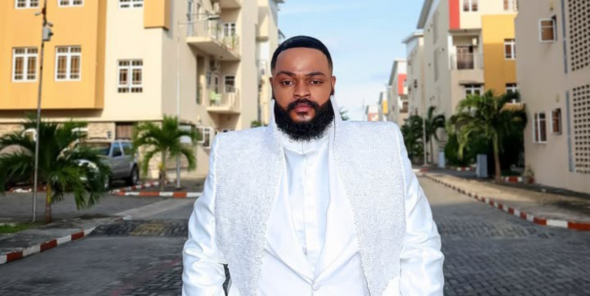 Man with a full beard in a white suit with a sparkling vest standing in a sunny residential street.