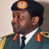 Profile view of a Black male military officer in ceremonial dress blues/greens, with gold shoulder insignia, red collar patches, and a black beret bearing an emblem, looking to the left.