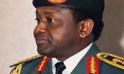 Profile view of a Black male military officer in ceremonial dress blues/greens, with gold shoulder insignia, red collar patches, and a black beret bearing an emblem, looking to the left.