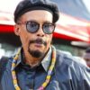 Portrait of a man wearing a black beret and sunglasses, with a colorful beaded necklace and a headset, outdoors at an event.”,