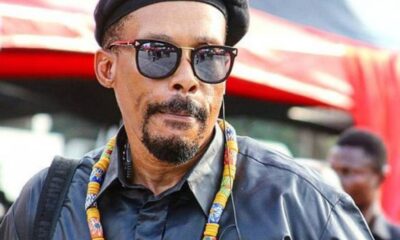 Portrait of a man wearing a black beret and sunglasses, with a colorful beaded necklace and a headset, outdoors at an event.”,