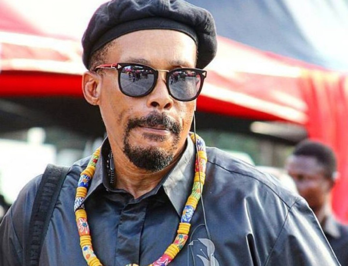 Portrait of a man wearing a black beret and sunglasses, with a colorful beaded necklace and a headset, outdoors at an event.”,