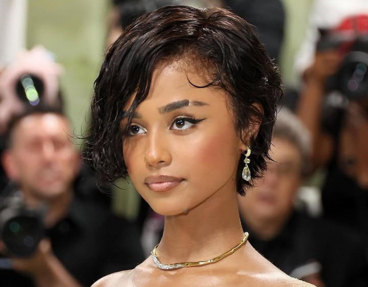 Young woman with short wet-look hair, diamond teardrop earrings, and a gold necklace at a red-carpet event.