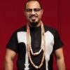Man with glasses in a black shirt with white pattern and long beaded necklaces, smiling against a red background.