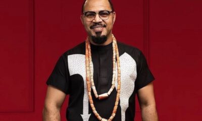 Man with glasses in a black shirt with white pattern and long beaded necklaces, smiling against a red background.
