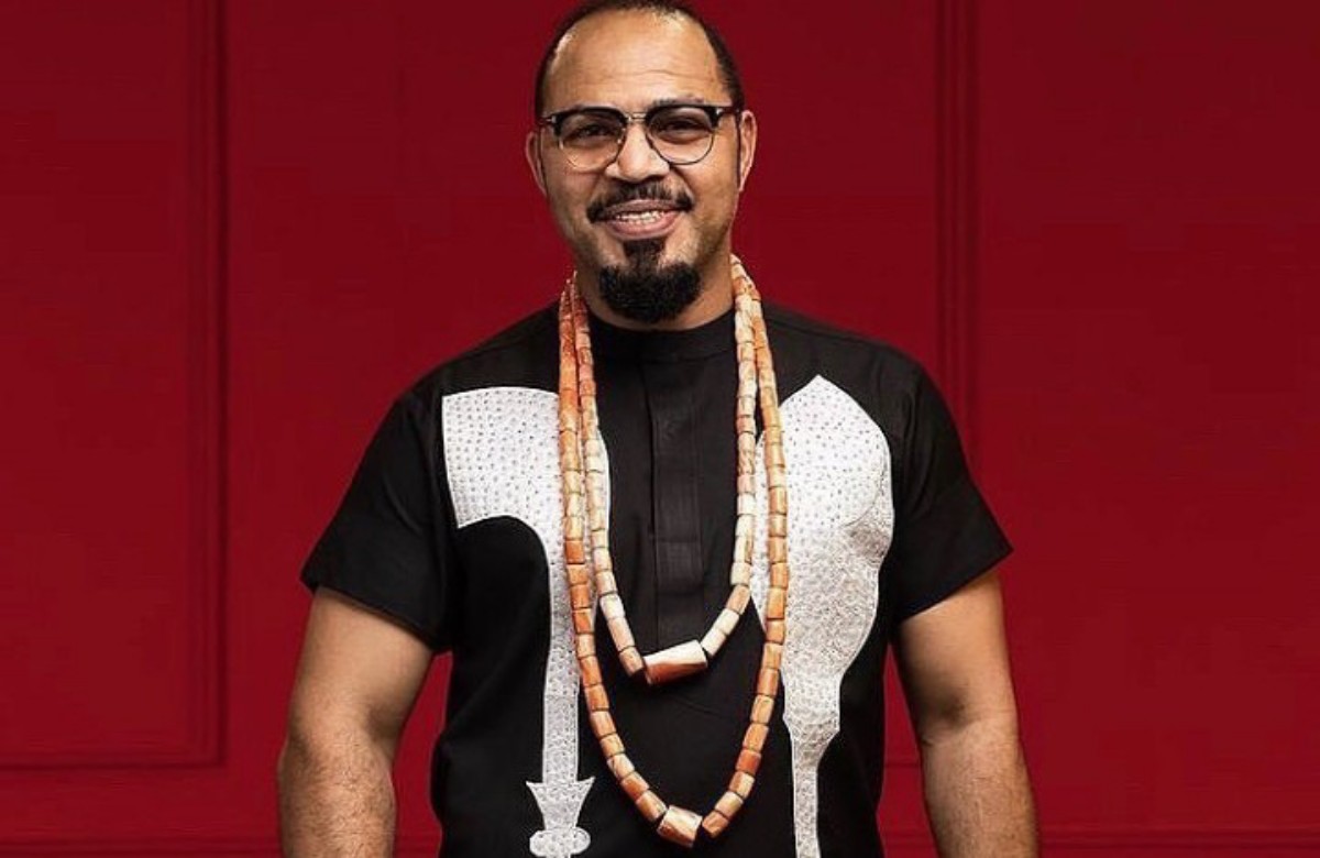 Man with glasses in a black shirt with white pattern and long beaded necklaces, smiling against a red background.