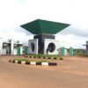 Gated entrance with a circular roundabout and modern concrete structures topped by a large green diamond-shaped canopy at the center.