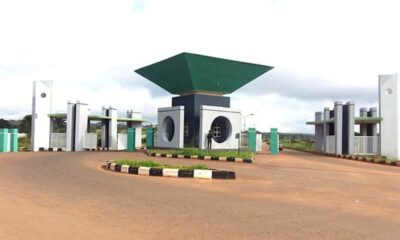 Gated entrance with a circular roundabout and modern concrete structures topped by a large green diamond-shaped canopy at the center.