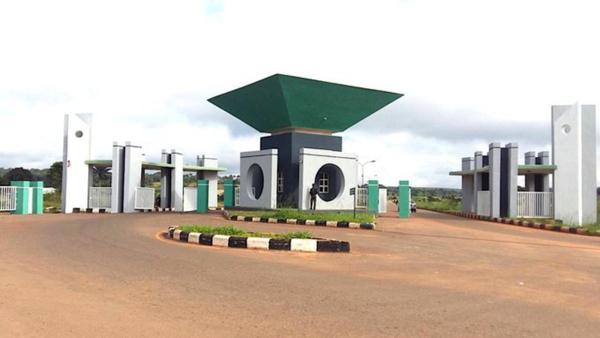 Gated entrance with a circular roundabout and modern concrete structures topped by a large green diamond-shaped canopy at the center.
