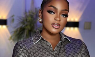 Close-up of a woman with braided hair and bold makeup wearing a patterned designer blouse in a softly lit room with warm and purple hues.