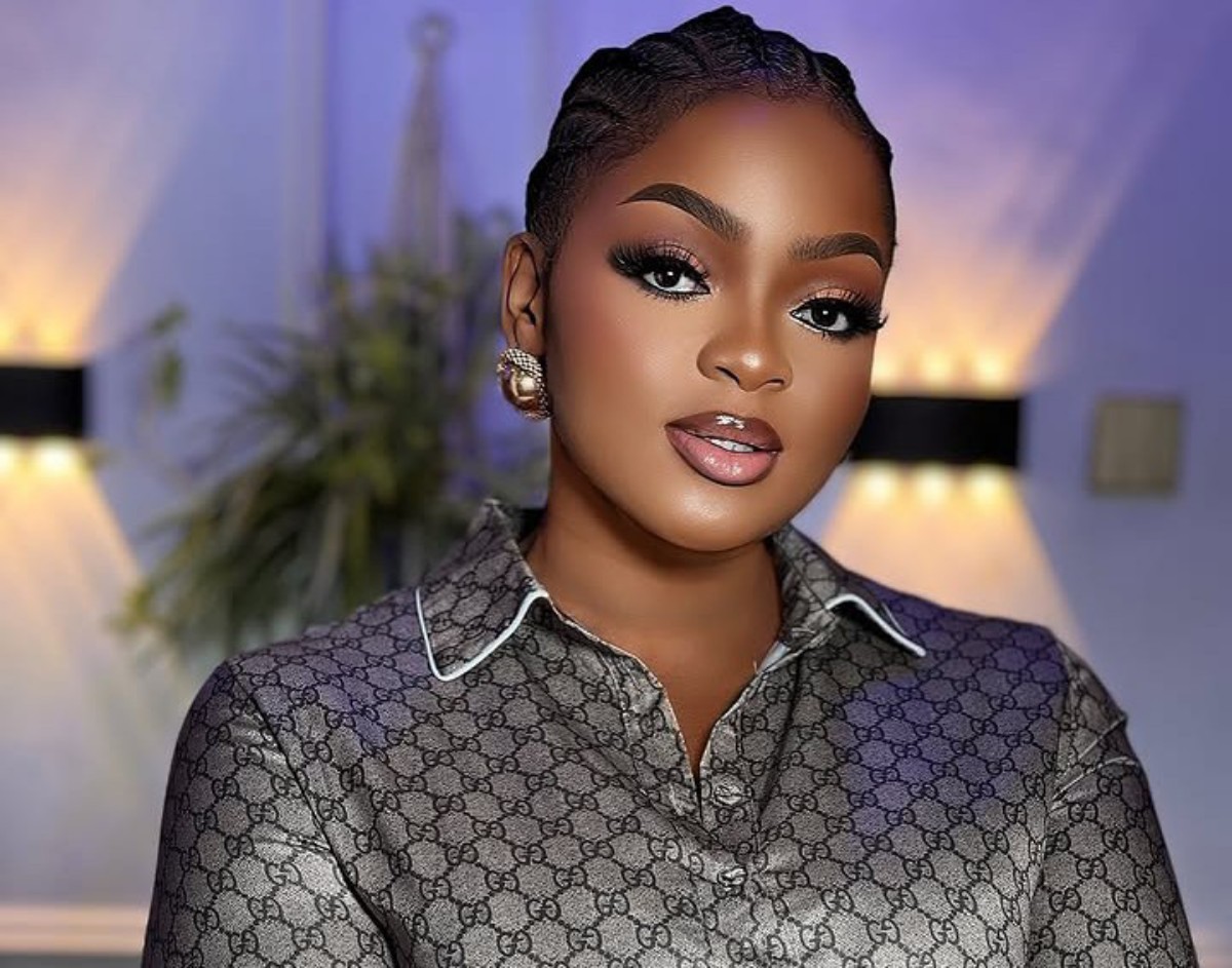 Close-up of a woman with braided hair and bold makeup wearing a patterned designer blouse in a softly lit room with warm and purple hues.