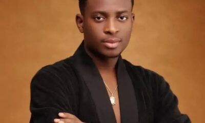 Portrait of a young man with short hair wearing a black velvet jacket, gold pendant, and crossed arms against a warm brown backdrop.