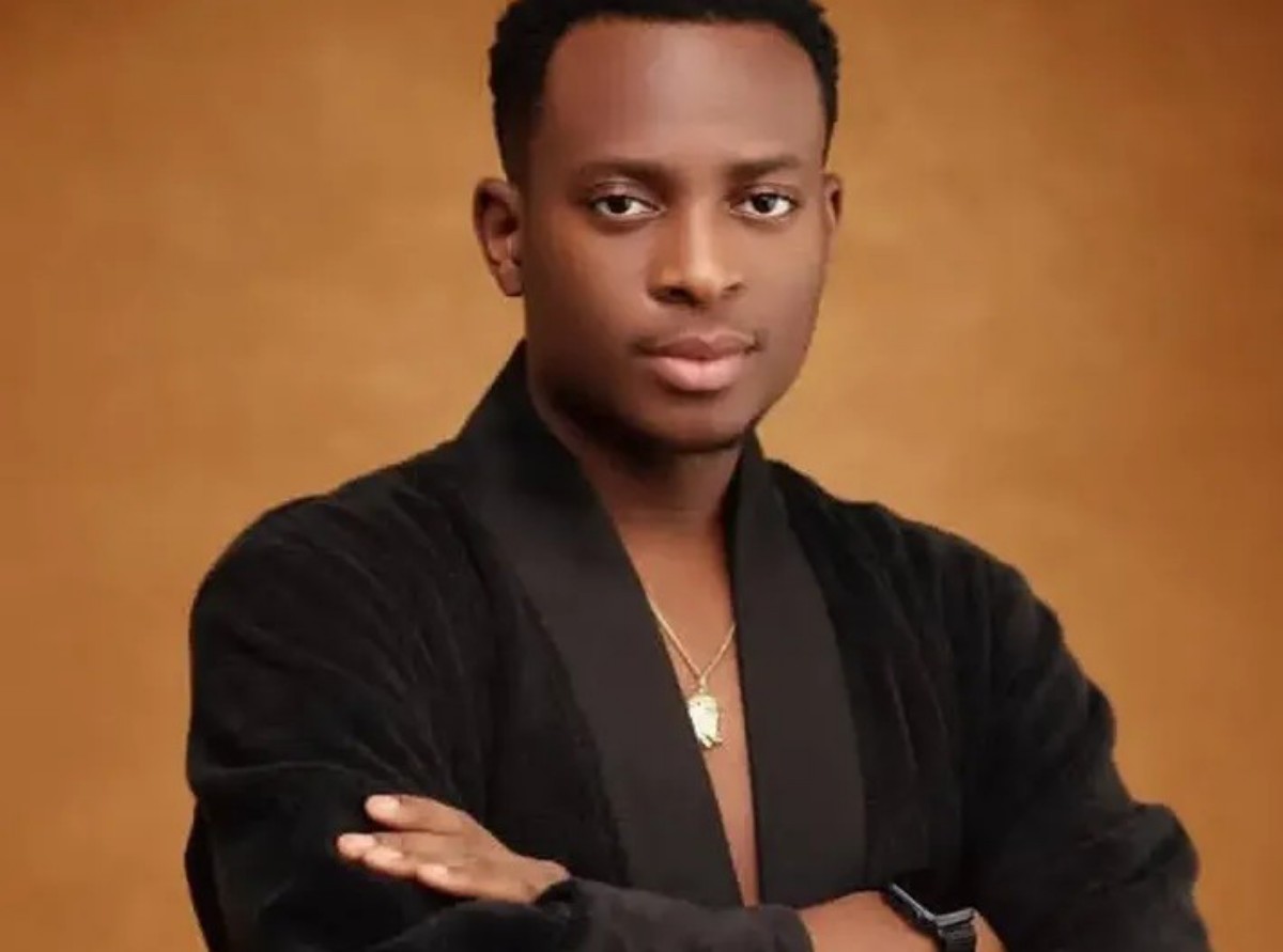 Portrait of a young man with short hair wearing a black velvet jacket, gold pendant, and crossed arms against a warm brown backdrop.