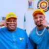 Two men in blue outfits and yellow caps pose together indoors, making hand signs with a Nigerian flag and emblems in the background.