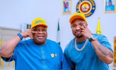 Two men in blue outfits and yellow caps pose together indoors, making hand signs with a Nigerian flag and emblems in the background.