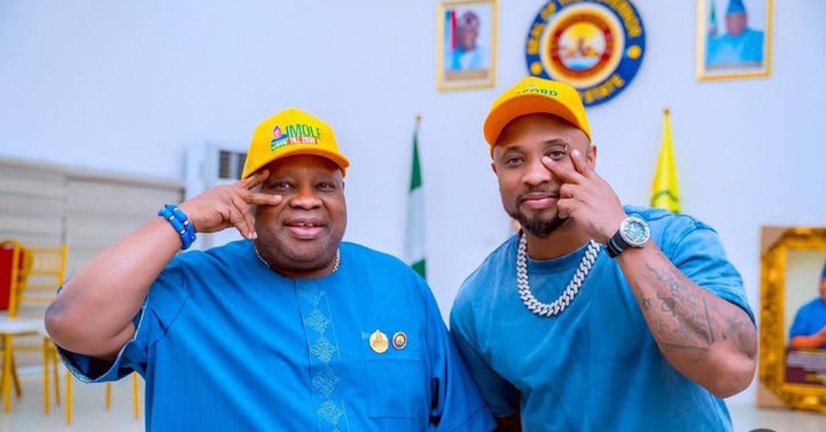 Two men in blue outfits and yellow caps pose together indoors, making hand signs with a Nigerian flag and emblems in the background.