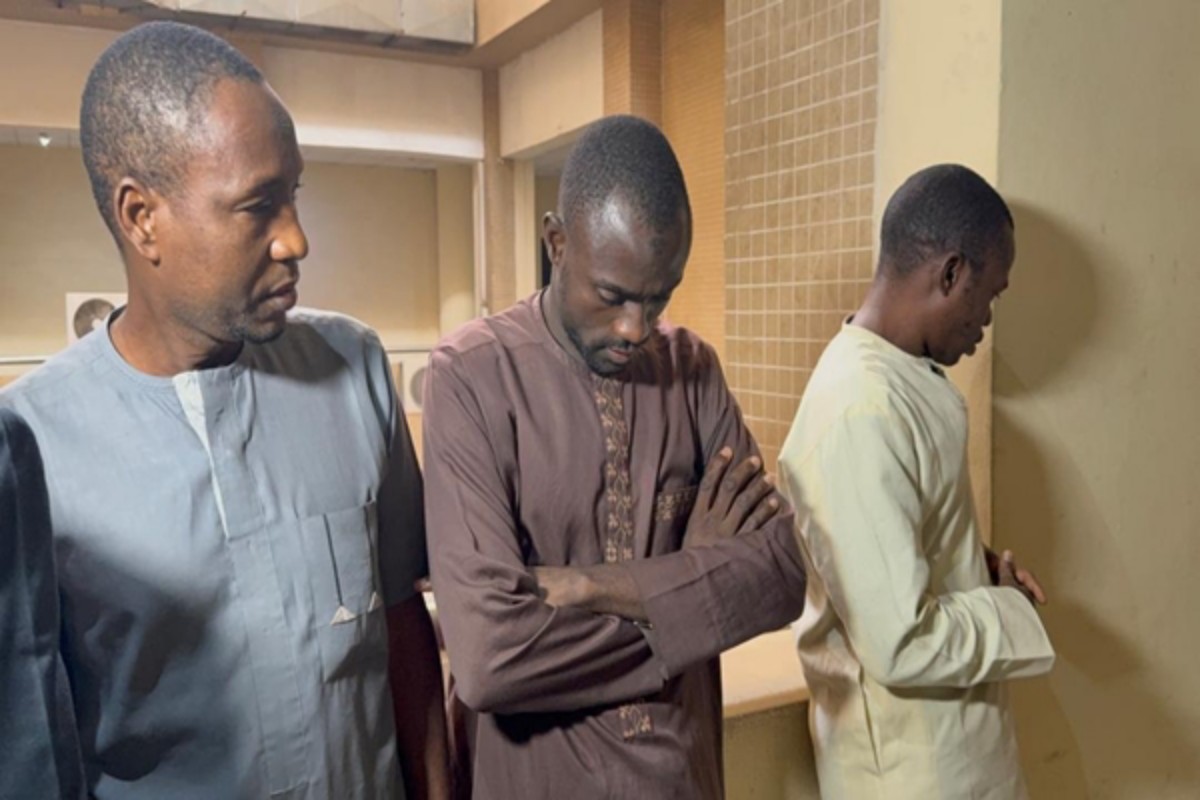 Three men in traditional clothing stand in a corridor, looking down with arms crossed or by their sides, appearing thoughtful or solemn.