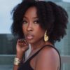 Portrait of a Black woman with natural curly hair, wearing a black sleeveless top and large gold earrings, looking at the camera.