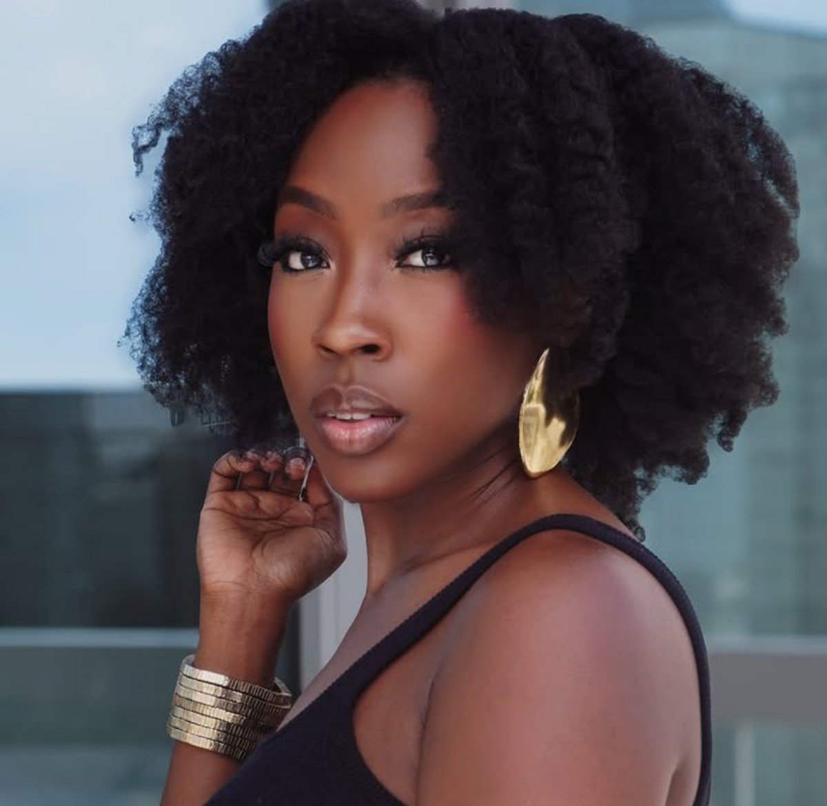 Portrait of a Black woman with natural curly hair, wearing a black sleeveless top and large gold earrings, looking at the camera.