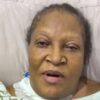 Older Black woman lying in bed propped on a pillow, mouth slightly open and eyes tired, wearing a light hospital gown and blanket nearby.
