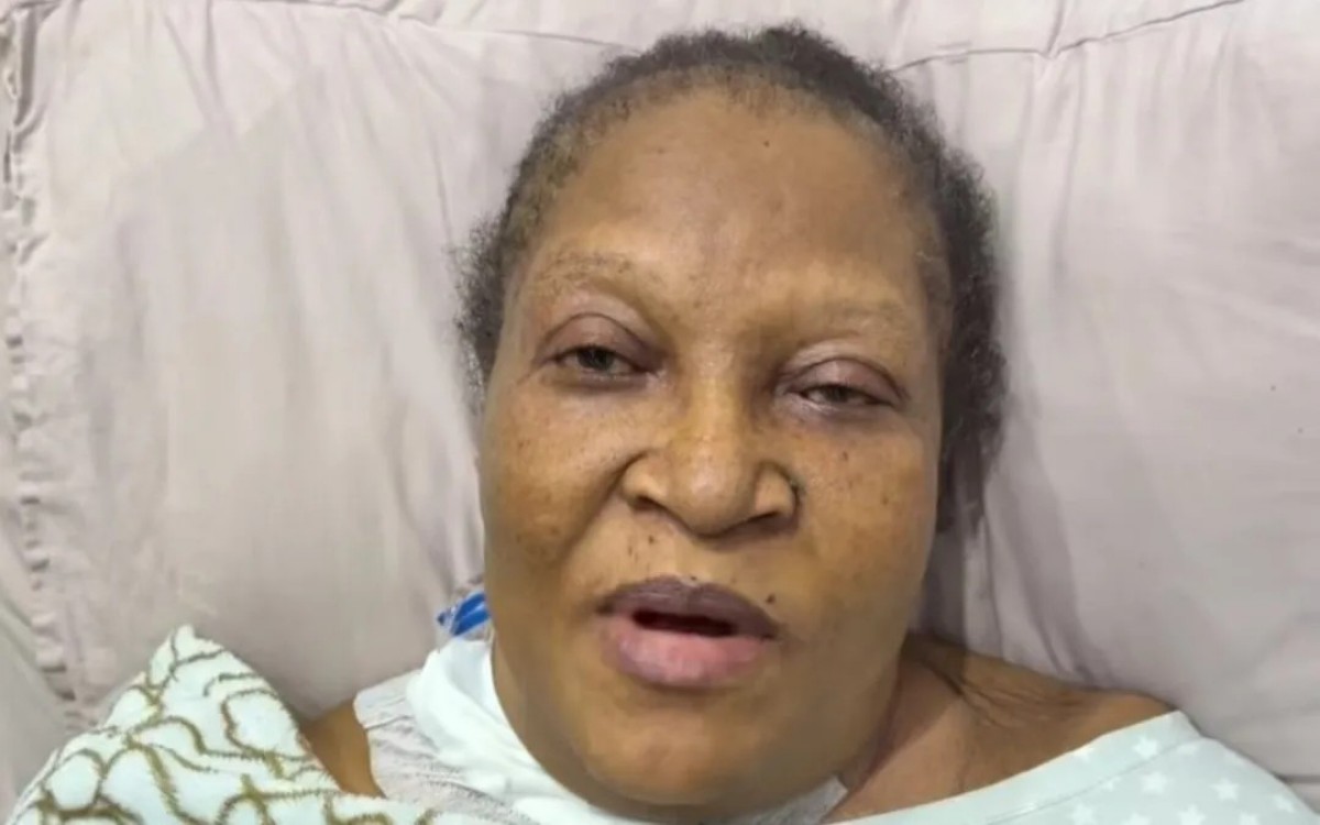 Older Black woman lying in bed propped on a pillow, mouth slightly open and eyes tired, wearing a light hospital gown and blanket nearby.