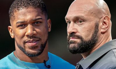 Two men standing side by side outdoors, the left wearing a blue sweater and a mic, the right bald with a beard, both looking ahead.