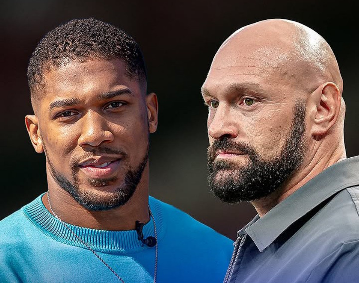 Two men standing side by side outdoors, the left wearing a blue sweater and a mic, the right bald with a beard, both looking ahead.
