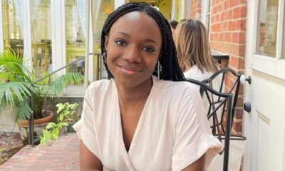 Smiling young woman with braided hair sits at an outdoor cafe table beside a brick wall and plants.