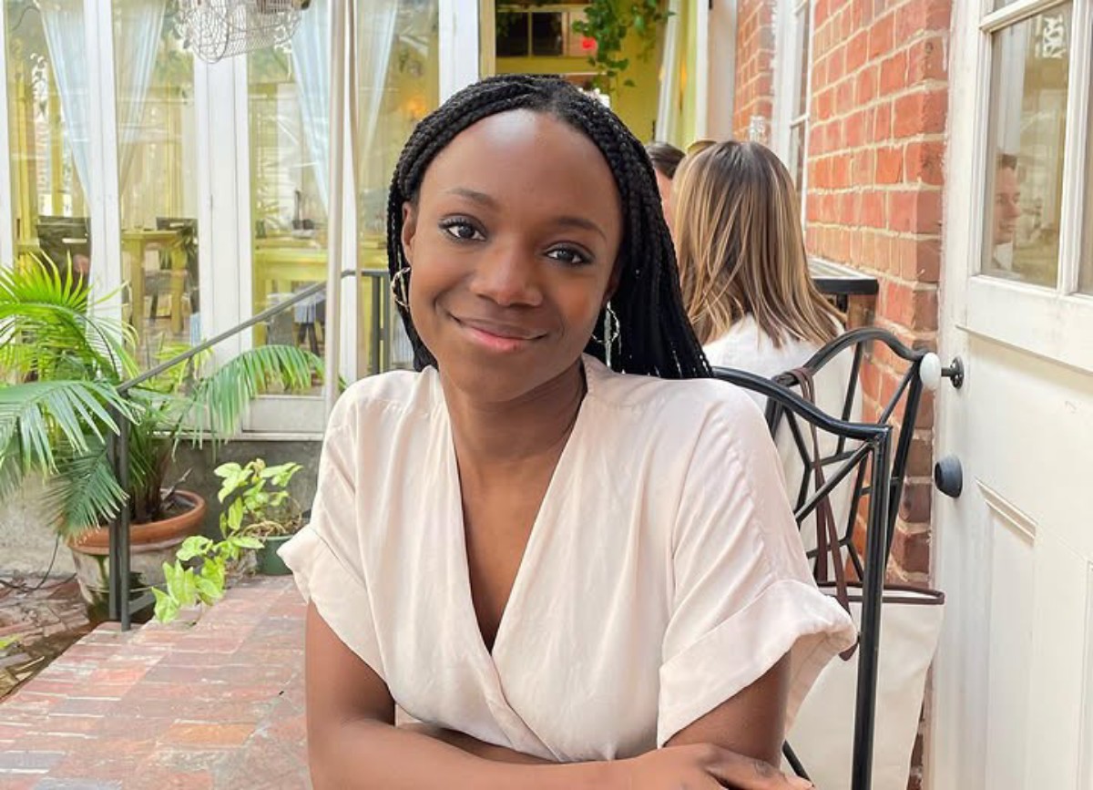 Smiling young woman with braided hair sits at an outdoor cafe table beside a brick wall and plants.