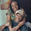 Two people share a warm hug in a home setting; a man in a black shirt wears a beige cap with 'FSG' while a smiling woman stands behind him in a colorful patterned top.