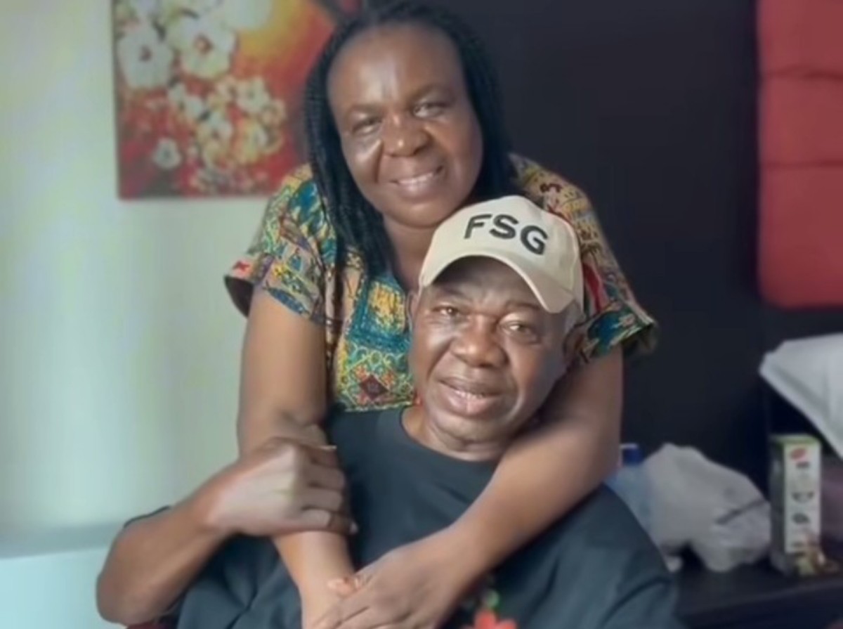 Two people share a warm hug in a home setting; a man in a black shirt wears a beige cap with 'FSG' while a smiling woman stands behind him in a colorful patterned top.