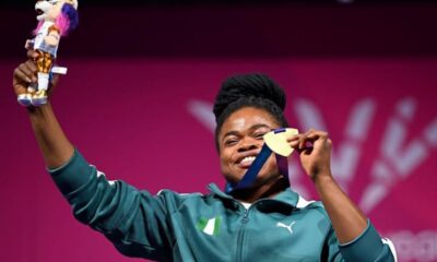 Athlete on a podium smiling, raising a gold medal and a trophy aloft against a pink background.