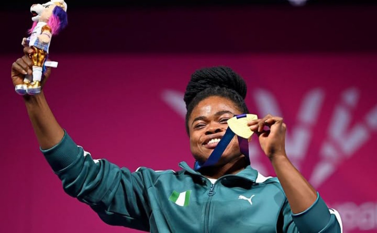 Athlete on a podium smiling, raising a gold medal and a trophy aloft against a pink background.