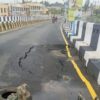 Motorcyclist rides past a large pothole and cracked asphalt on a barrier-lined road bridge/section.