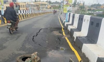 Motorcyclist rides past a large pothole and cracked asphalt on a barrier-lined road bridge/section.
