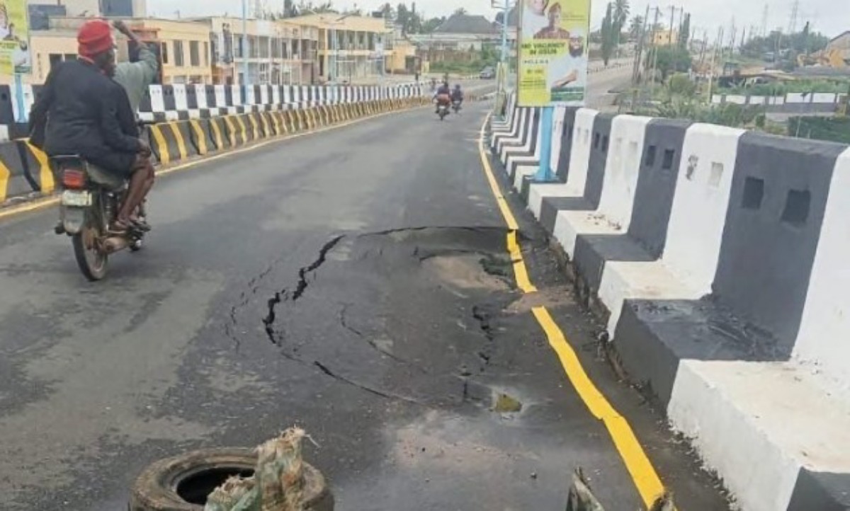 Motorcyclist rides past a large pothole and cracked asphalt on a barrier-lined road bridge/section.