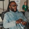 Man with long dreadlocks and a beard wearing oversized sunglasses, a light denim jacket, and jewelry sits in a chair, looking to the side.