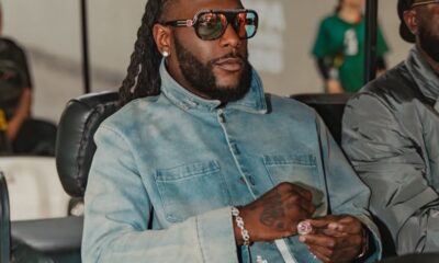 Man with long dreadlocks and a beard wearing oversized sunglasses, a light denim jacket, and jewelry sits in a chair, looking to the side.