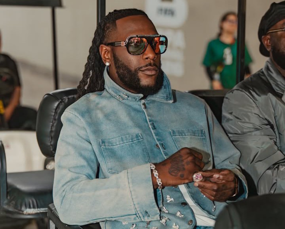 Man with long dreadlocks and a beard wearing oversized sunglasses, a light denim jacket, and jewelry sits in a chair, looking to the side.