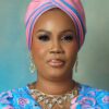 Portrait of a woman wearing a pink and blue head wrap, blue patterned dress, and gold jewelry against a soft teal background.