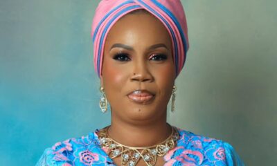 Portrait of a woman wearing a pink and blue head wrap, blue patterned dress, and gold jewelry against a soft teal background.