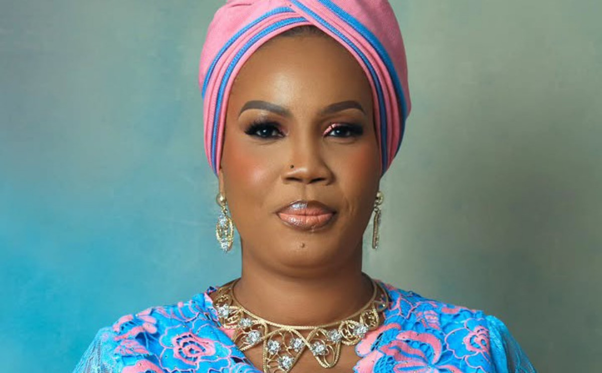 Portrait of a woman wearing a pink and blue head wrap, blue patterned dress, and gold jewelry against a soft teal background.