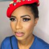 Close-up portrait of a woman with dramatic makeup, bold brows, long lashes, and bronze cheeks, wearing a red jeweled headwrap and blue top.