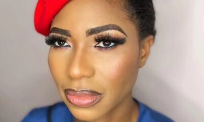 Close-up portrait of a woman with dramatic makeup, bold brows, long lashes, and bronze cheeks, wearing a red jeweled headwrap and blue top.