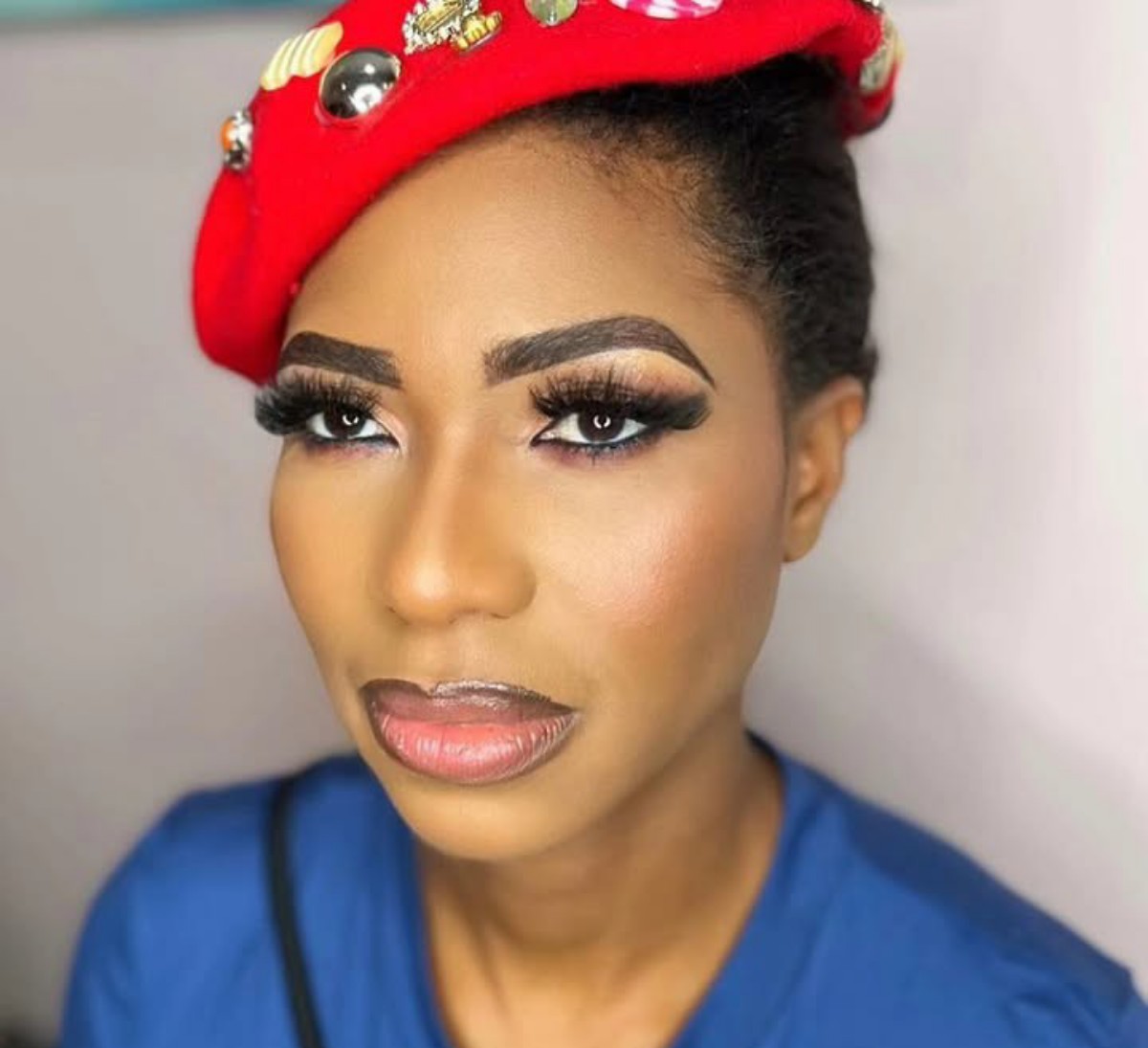 Close-up portrait of a woman with dramatic makeup, bold brows, long lashes, and bronze cheeks, wearing a red jeweled headwrap and blue top.