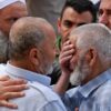 Two older men in light shirts share a comforting embrace as others surround them in a tense moment.