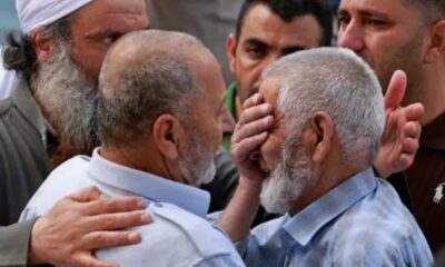 Two older men in light shirts share a comforting embrace as others surround them in a tense moment.